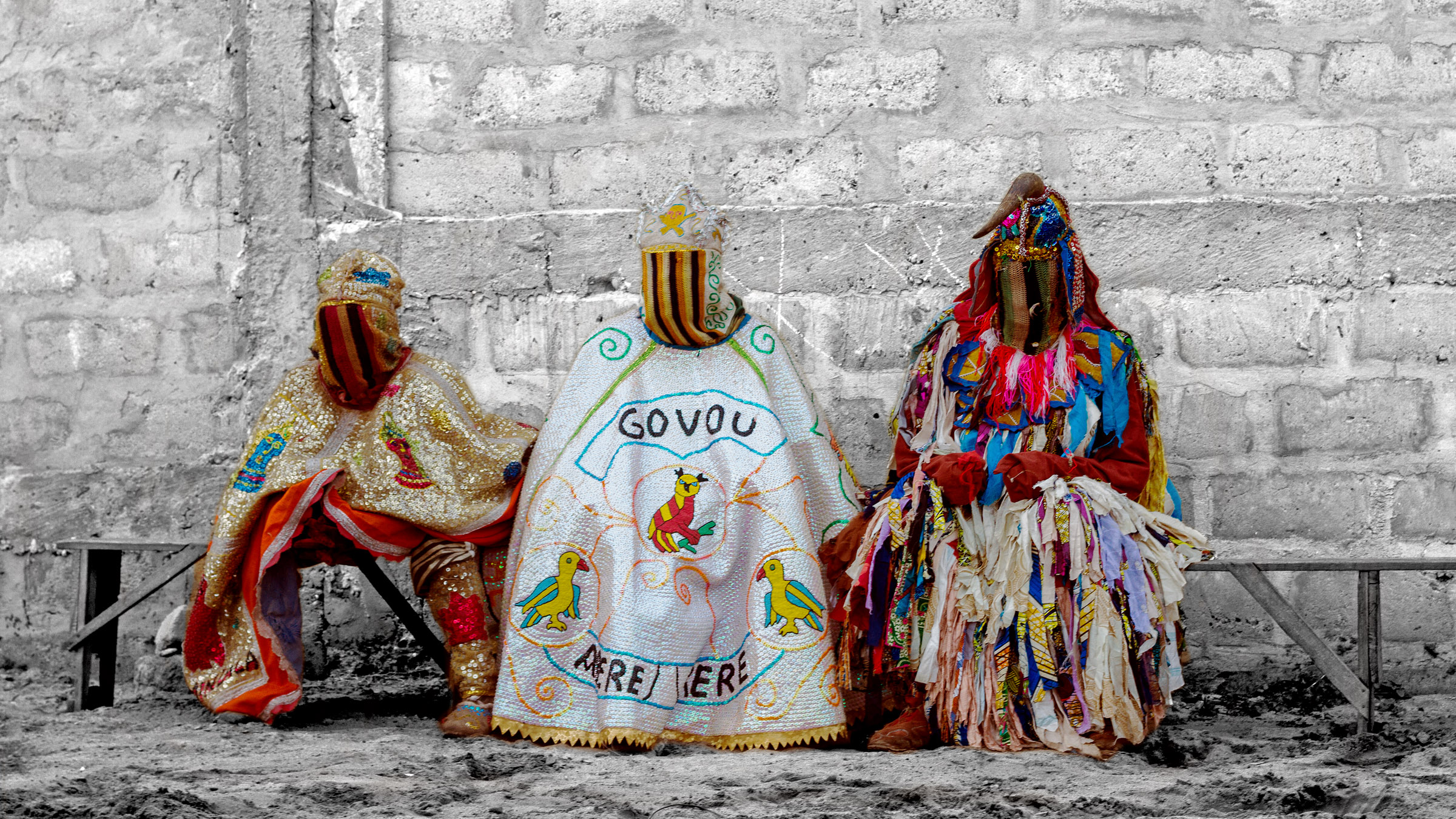 Three Egungun masquerade performers sit side by side on a bench